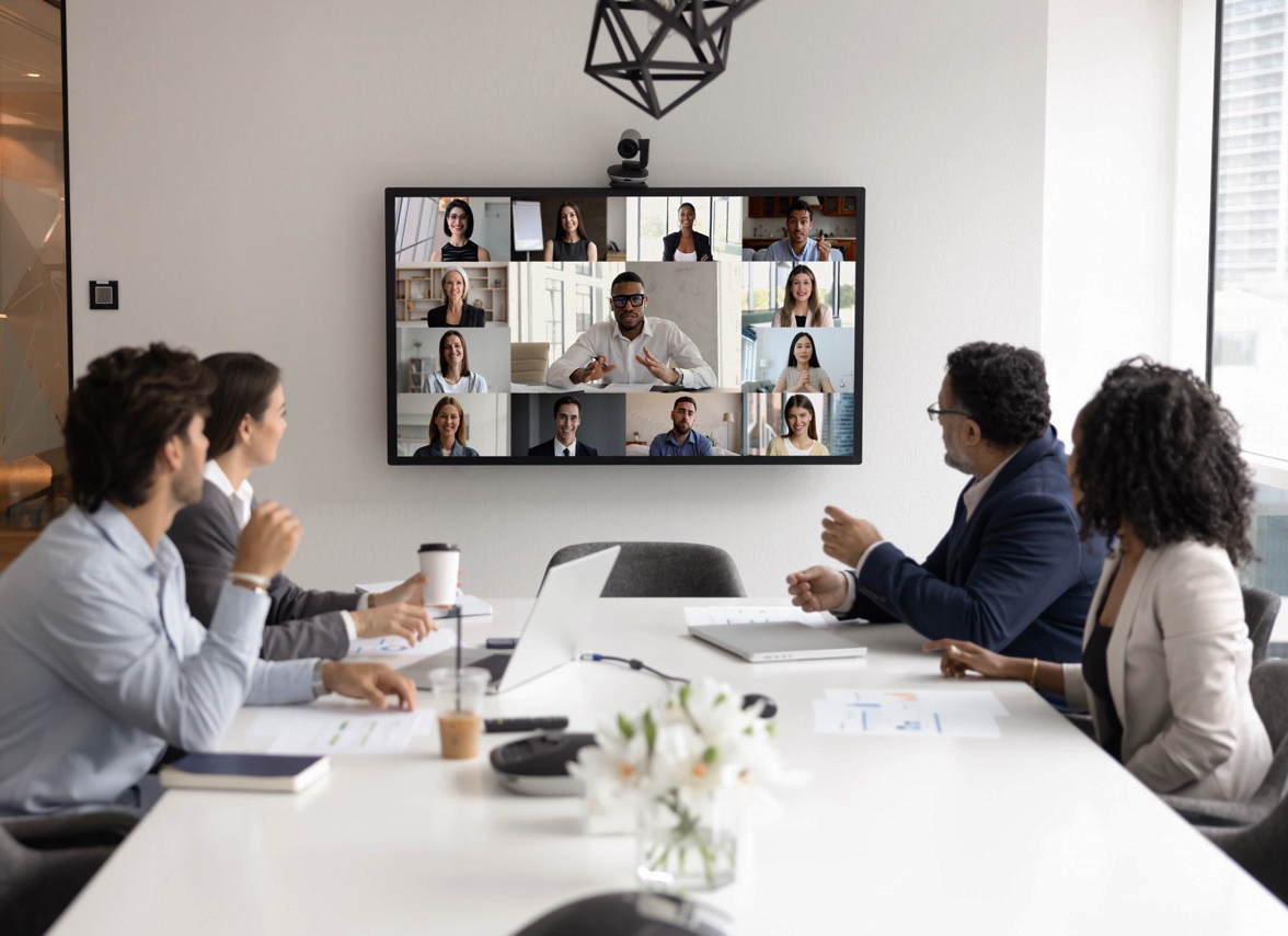 Four business professionals sit around a white board table. A screen on the wall projects the faces of their colleagues as they take part in a virtual meeting.