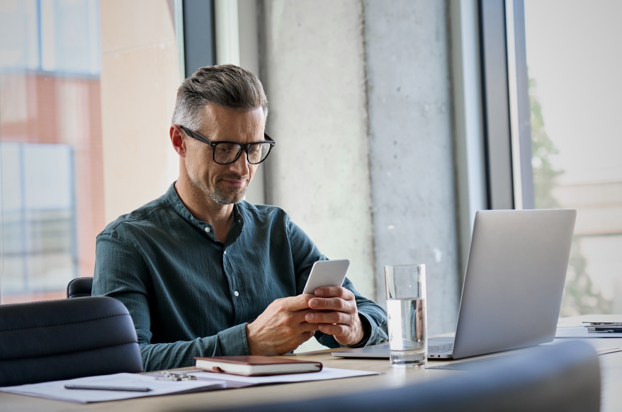 A business man smiles as he looks down at his phone while sitting at his desk. A laptop is open in front of him.