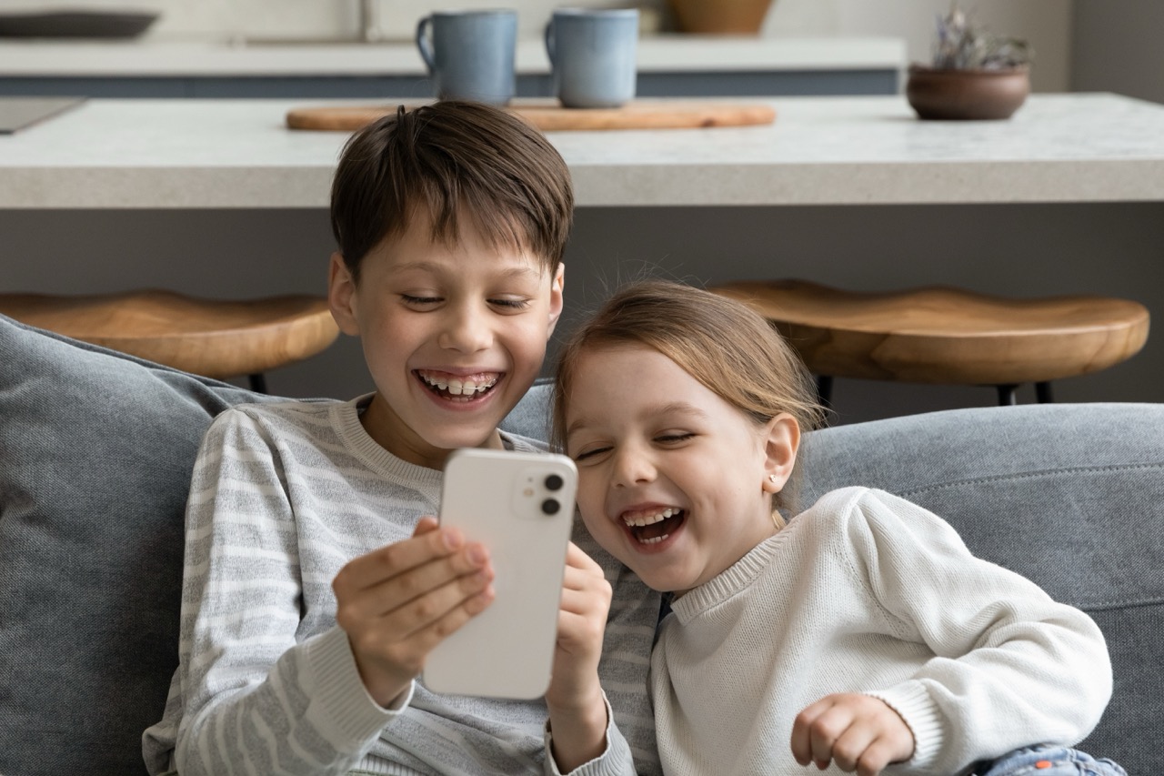 A young brother and sister laugh as they watch something on a phone.