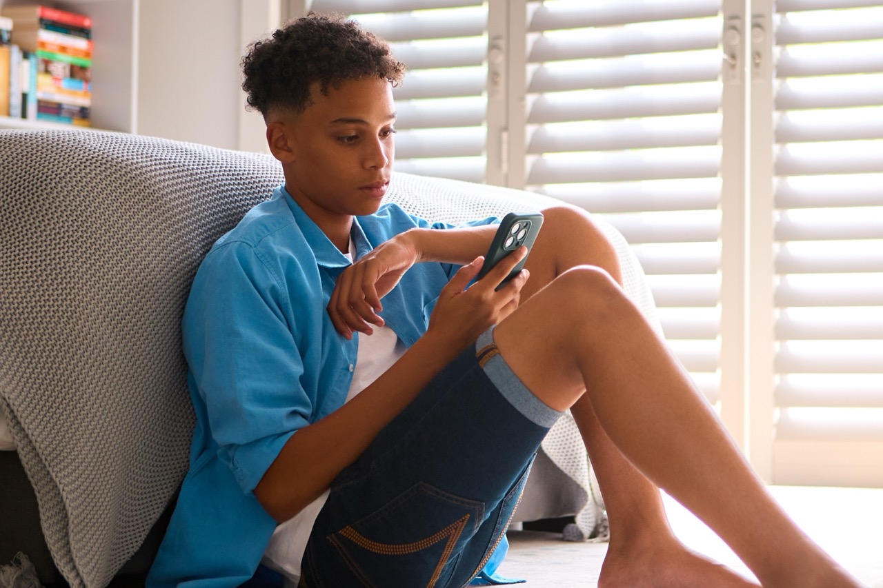 A teenage boy sits on the floor of his bedroom and leans against his bed as he watches something on his phone.