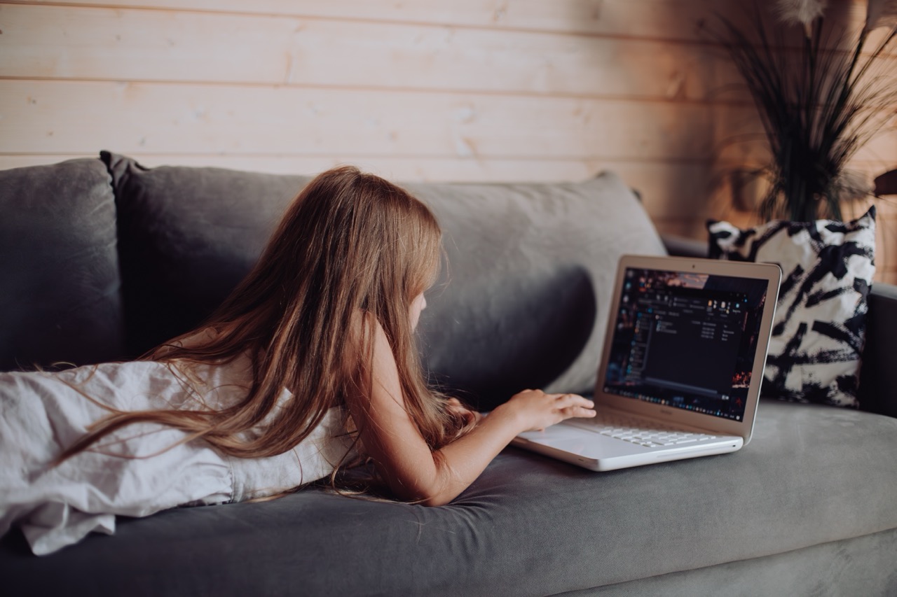 A young girl lies on her stomach on a sofa, a laptop open in front of her.