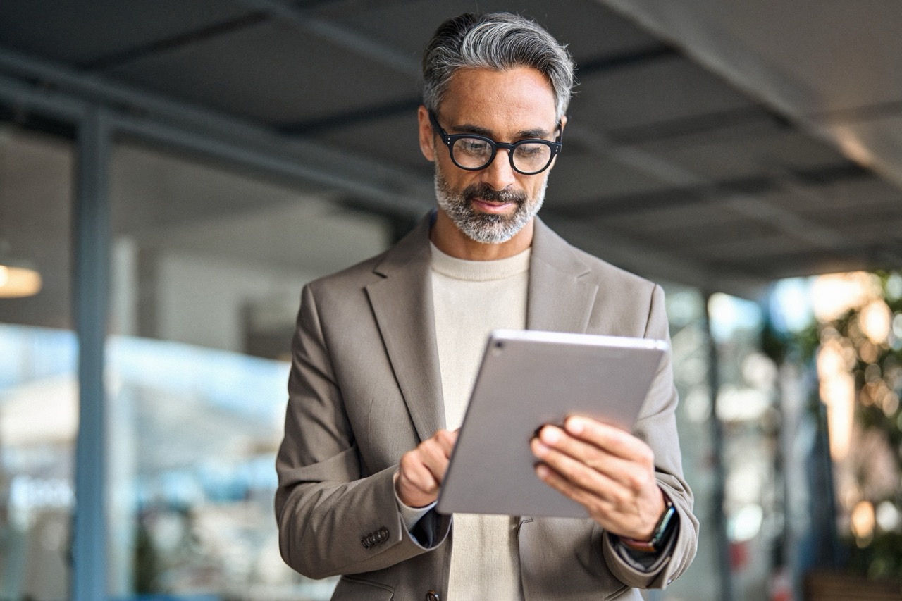 Older business man wearing a grey blazer and trendy glasses smiles as he looks down at his tablet. 