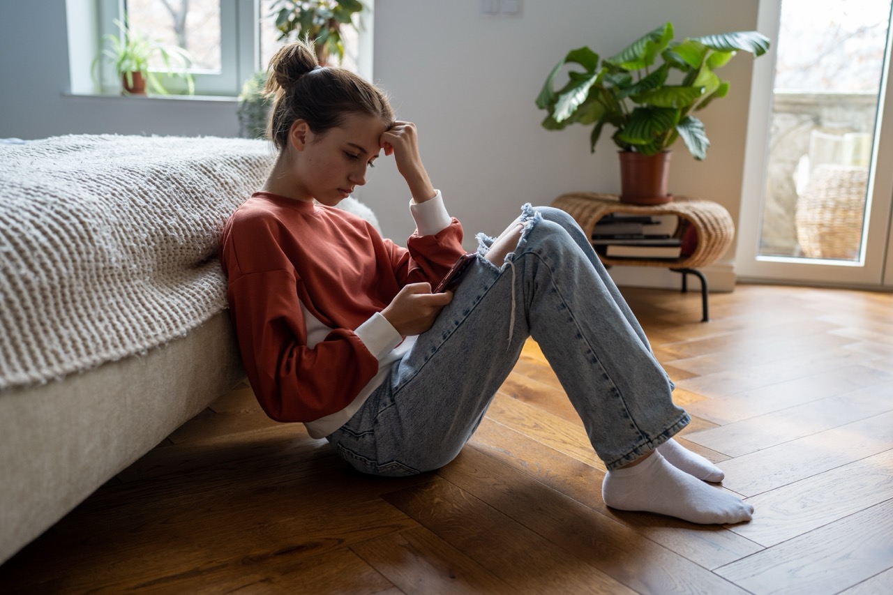 A teen girl looks sad as she stares down at her phone while sitting on the floor in her bedroom.