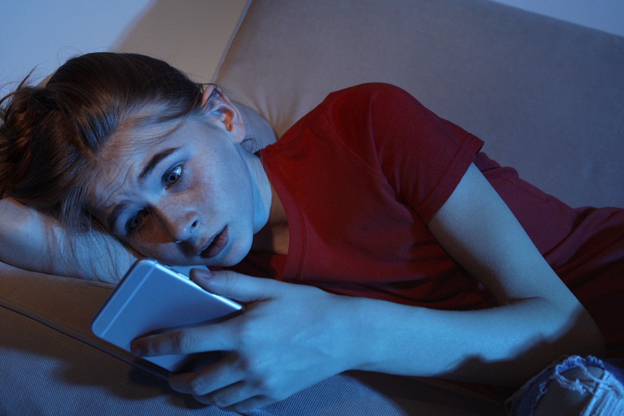 Shocked teenage girl with smartphone on sofa in dark room.
